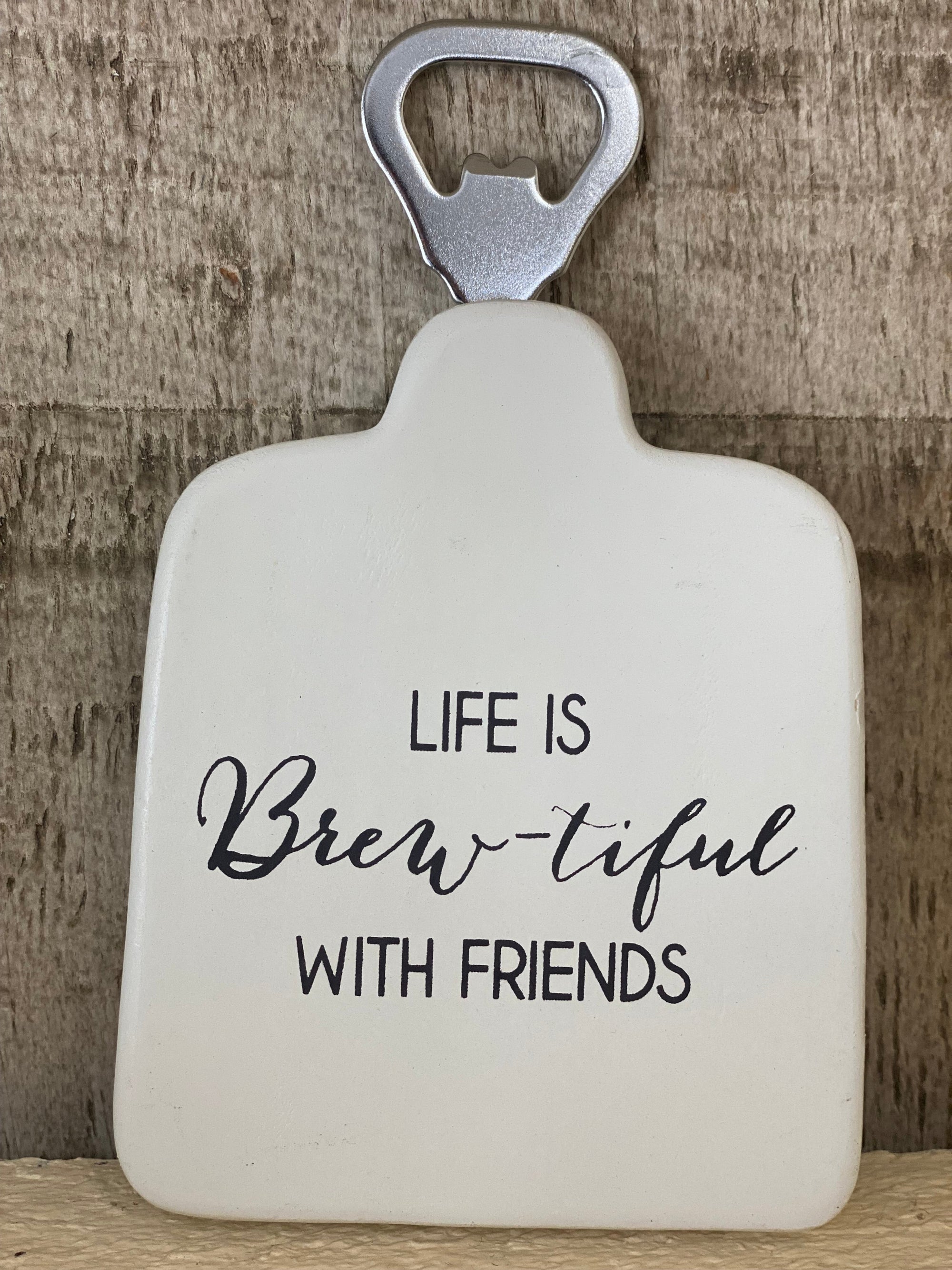 A white ceramic bottle opener with the phrase 'LIFE IS Brew-tiful WITH FRIENDS' printed in black, set against a rustic wood background. Set of 4 with storage box. Renfroe Pecan Co. Pensacola, FL