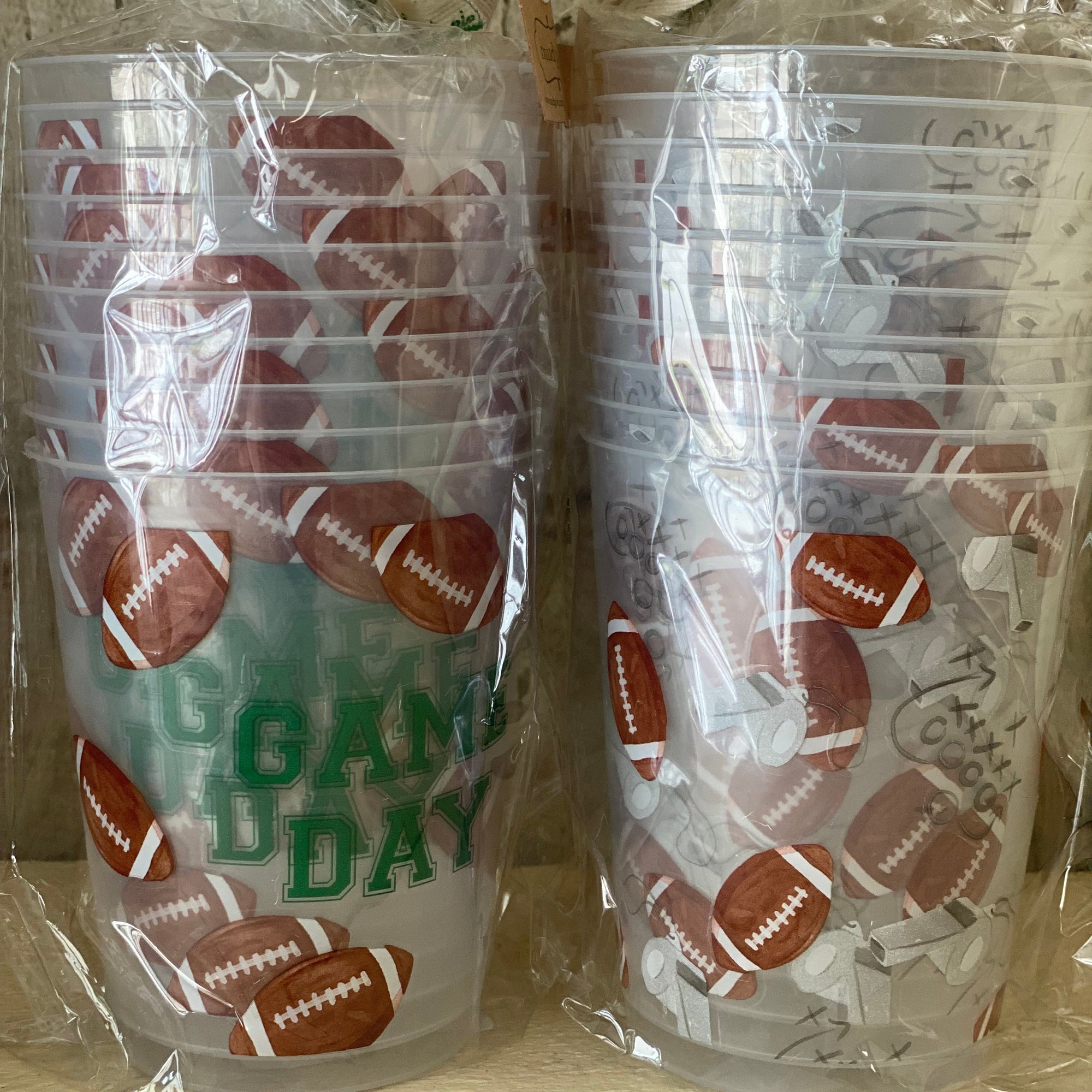 Two stacks of clear plastic cups wrapped in clear cellophane and tied with white ribbon handles, printed with brown footballs and green 'GAME DAY' text, on a light wood surface. Renfroe Pecan Co. Pensacola, FL