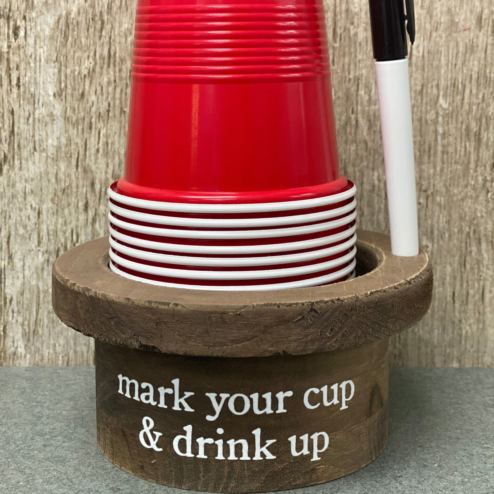 A rustic wooden cup holder and desk organizer with the phrase 'mark your cup & drink up' printed on the front. The holder contains a stack of red plastic cups and a black-and-white marker, set against a light wood panel background. Renfroe Pecan Co. Pensacola, FL