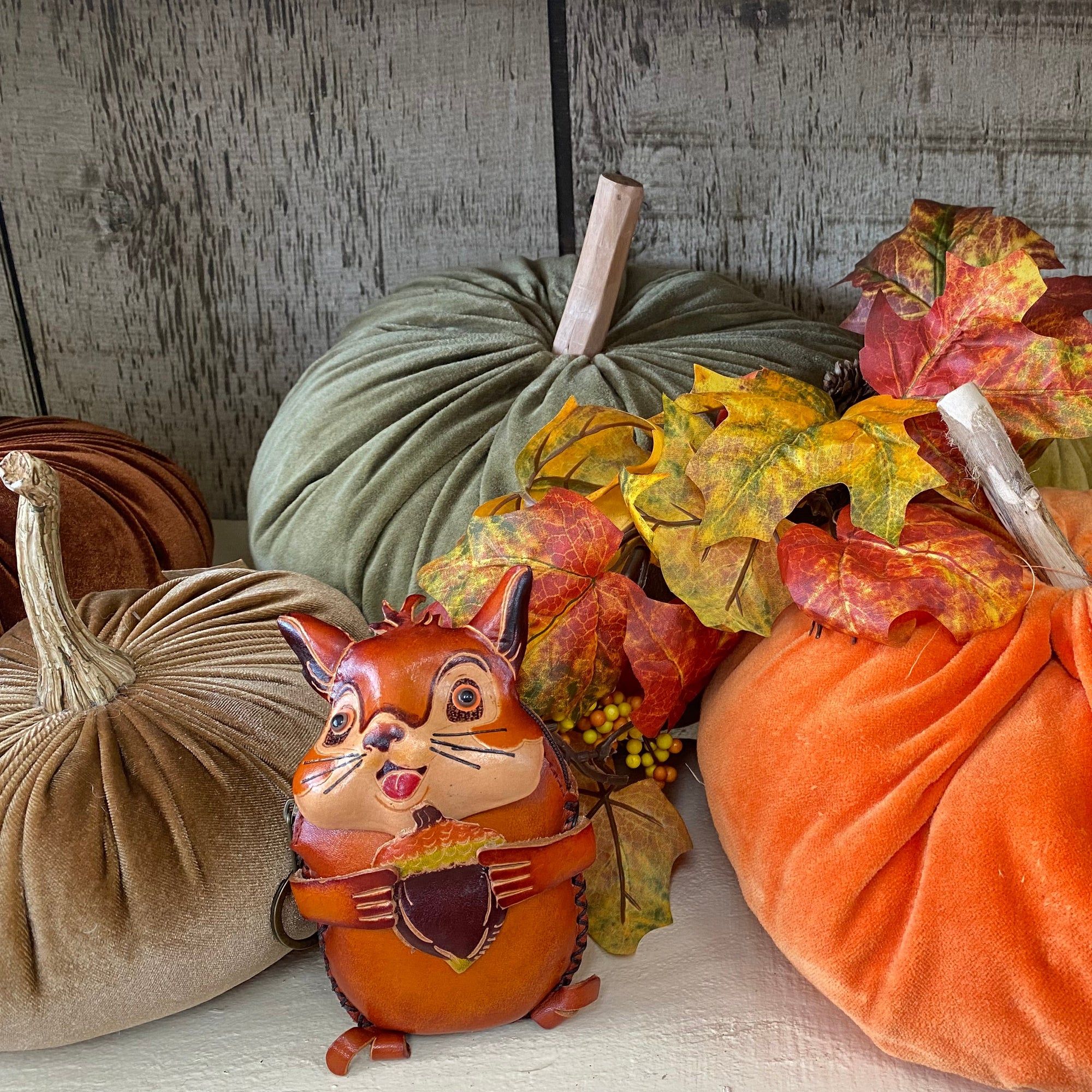 A collection of autumn-themed velvet decorations including pumpkins in orange, sage green, and beige, styled with faux fall leaves and two ceramic squirrel figurines holding acorns on a white shelf against a rustic gray wood plank background. Renfroe Pecan Co. Pensacola, FL