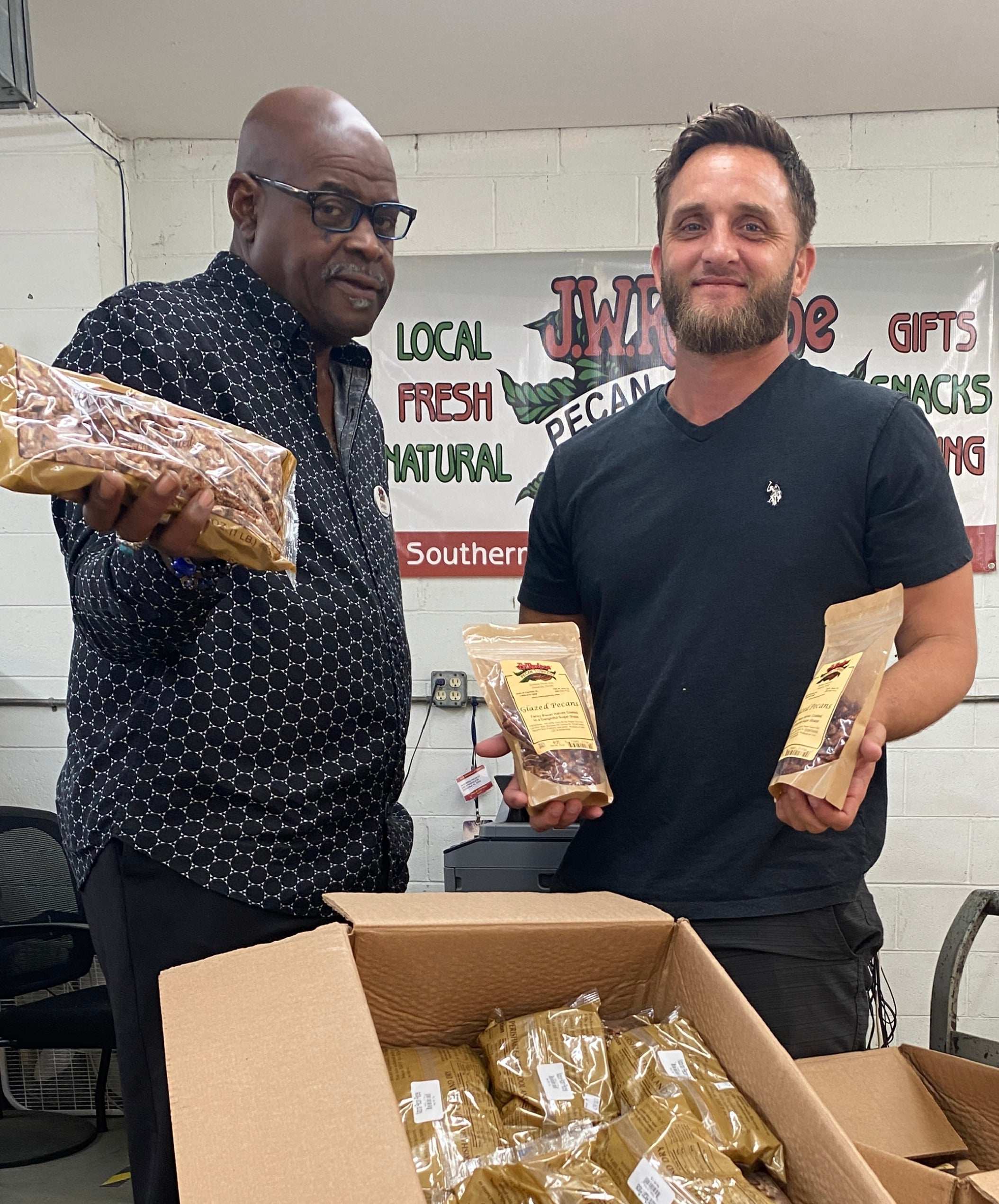 Two sales representatives holding Renfroe Pecan products in front of a box with more packages, in a setting with a sign in the background.