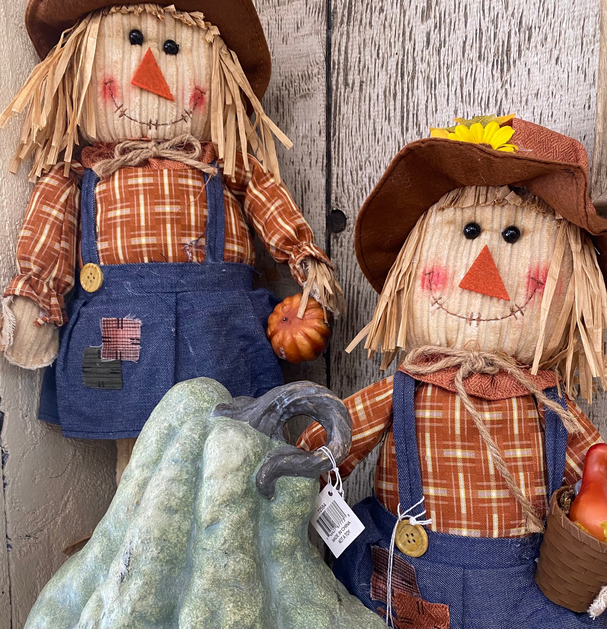 Two autumn scarecrow garden decorations with straw hair, brown hats, orange plaid shirts, blue denim-style overalls, and small pumpkins, posed against a weathered wood background. Renfroe Pecan Co. - Pensacola, FL